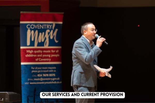 Image of Mark Steele talking on a microphone next to Coventry Music banner